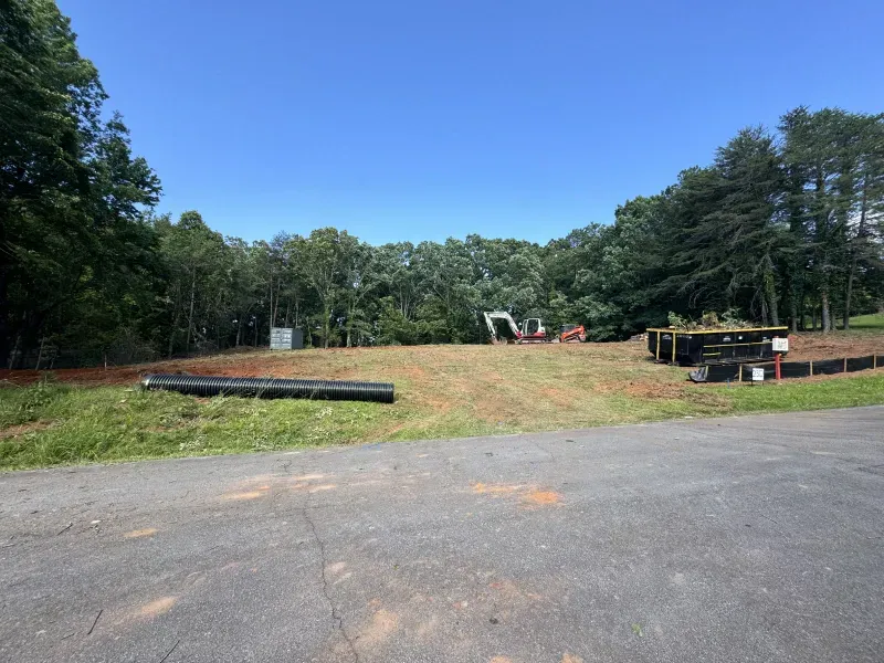 Construction site with cleared land, an excavator, and a black pipe. Trees and blue sky in background.