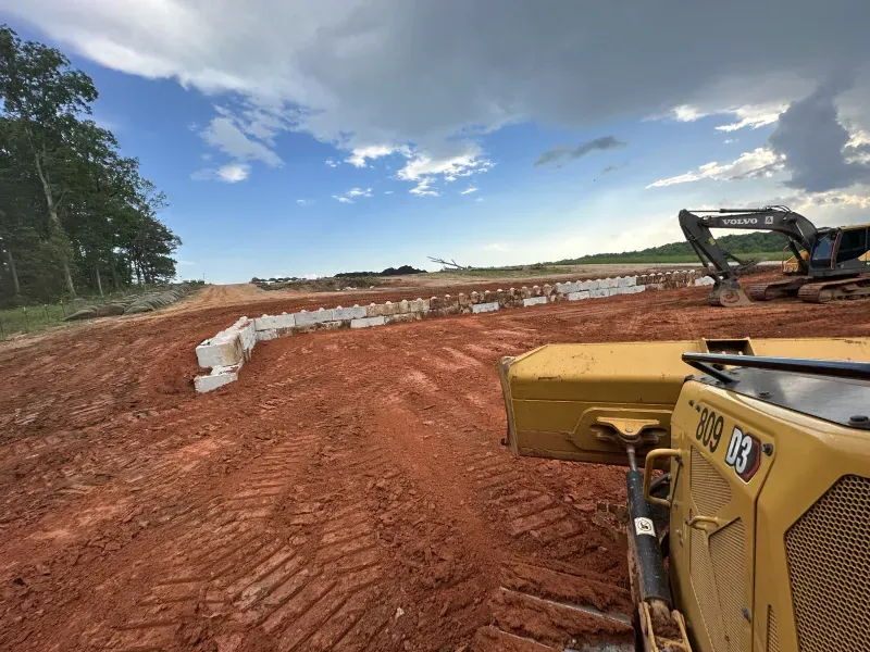 Construction site with bulldozer in foreground, earth movers, barriers, and cloudy sky.