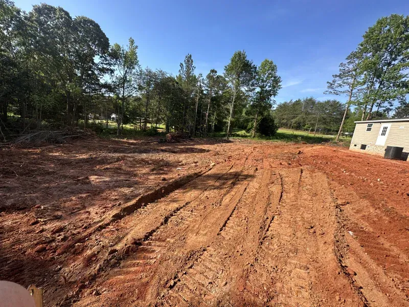 Cleared red dirt lot with tire tracks, trees in the background, blue sky, and a small building to the right.