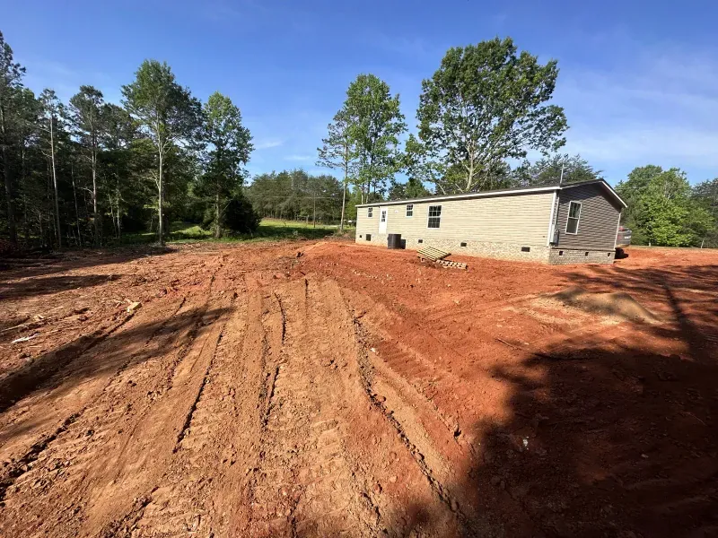 Dirt lot with a mobile home in a rural setting. Red dirt and tire tracks in foreground, trees in the background.