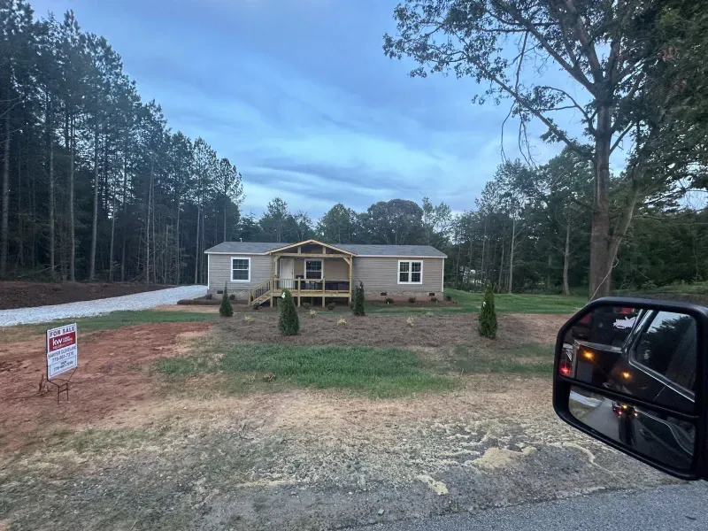 Tan single-story house with a small porch, set among trees and a dirt driveway. A sign is in the front yard.