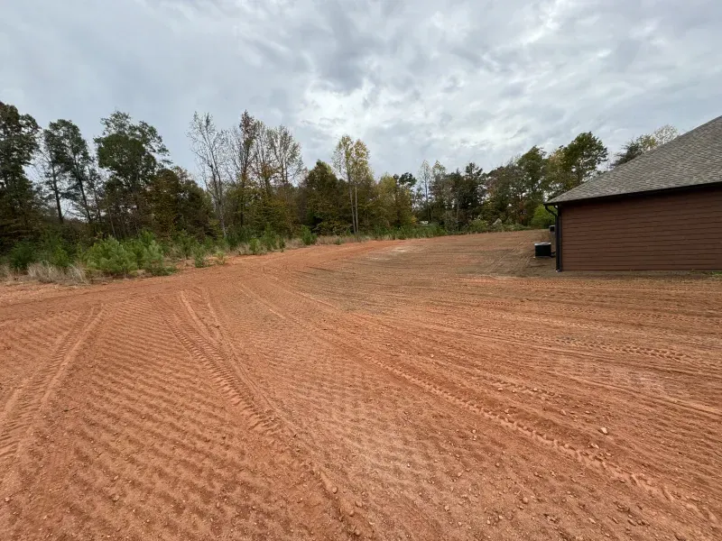Red dirt lot with tire tracks, trees in the background, and a brown building on the right under a cloudy sky.