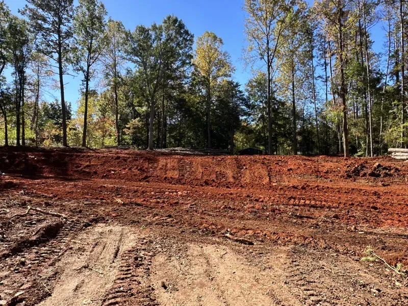 Dirt lot with red soil and tire tracks, cleared area near a tree line under a blue sky.