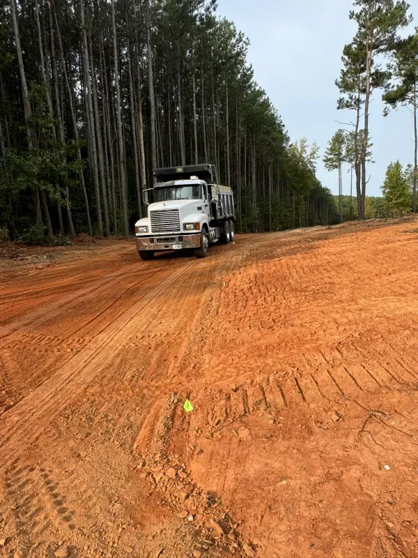 White dump truck on a dirt road in a forest. Red dirt, tall trees, cloudy sky.