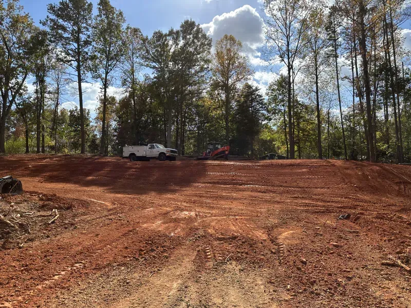 Cleared red dirt lot with work trucks and trees in the background. Sunny day.