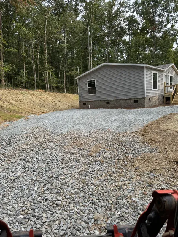 Gravel driveway leading to a gray house, with a wooded area in the background and a tractor in the foreground.