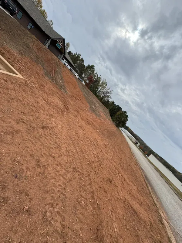 Red dirt ground sloped towards a road and water under a cloudy sky with a building and trees visible.