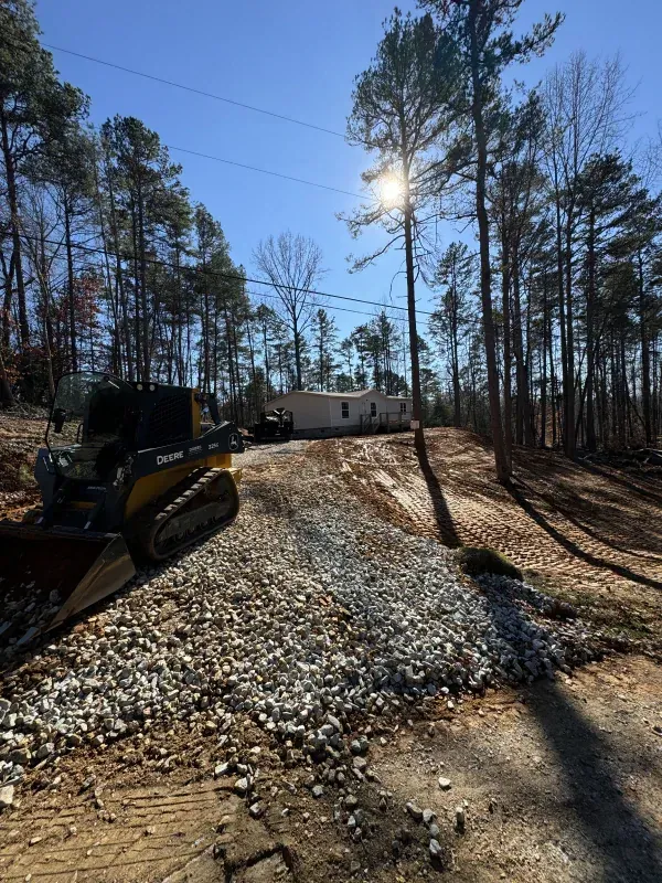 A compact track loader grading a gravel driveway leading to a house in a wooded area; sunny day.