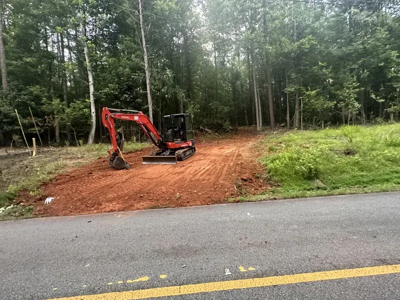 Mini excavator on dirt clearing next to a road, trees in the background.