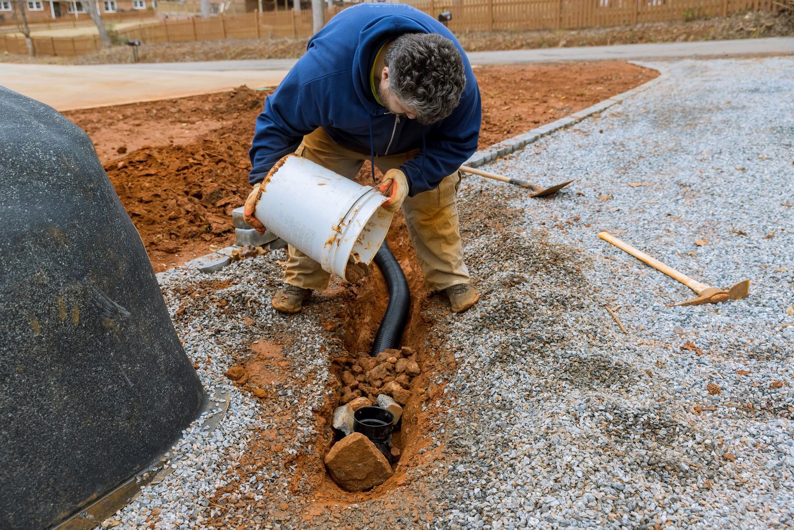 Man pouring from a bucket into a trench with a pipe, surrounded by gravel and dirt.