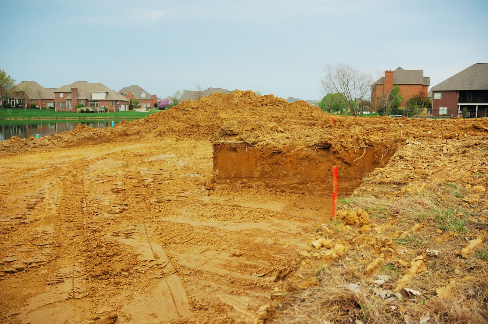 Construction site with dirt piles and a trench in front of residential houses and a lake.