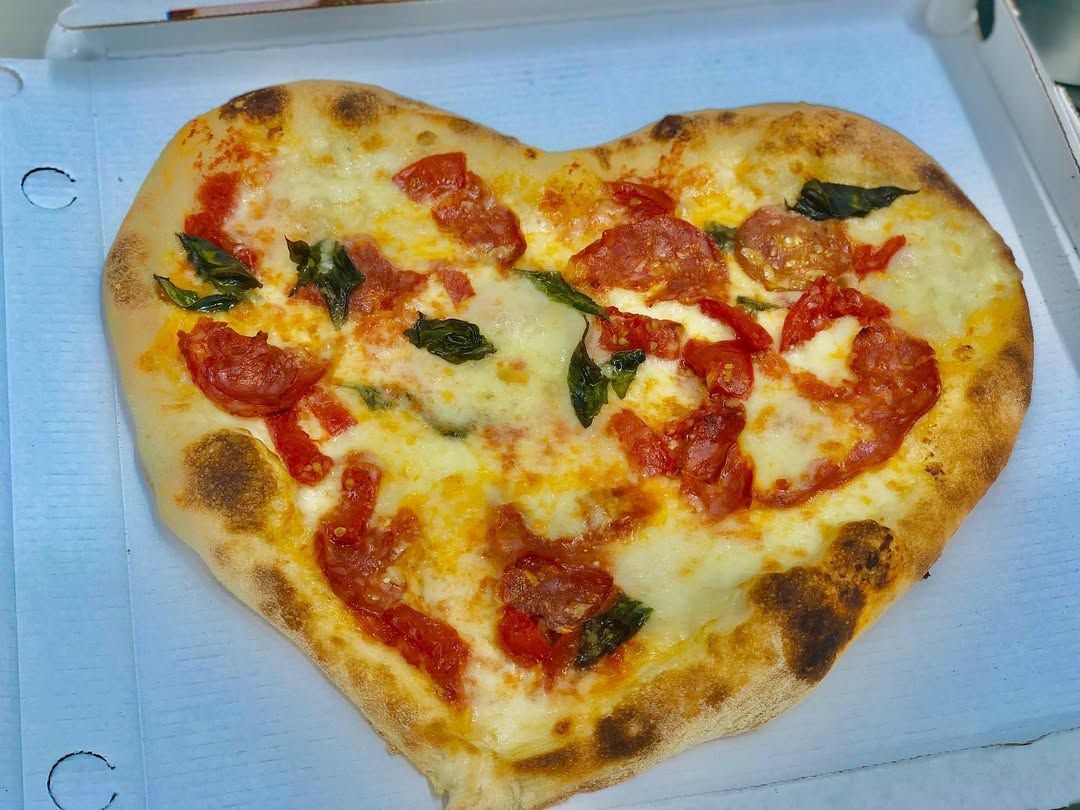 Pizza a forma di cuore in una scatola bianca, condita con salsa di pomodoro, formaggio fuso e foglie di basilico.