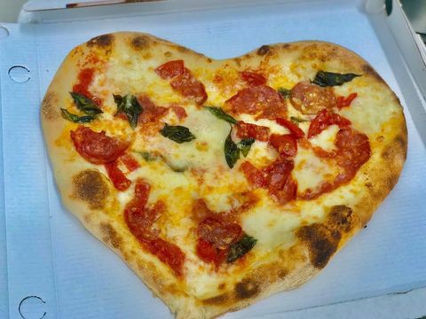 Pizza a forma di cuore in una scatola bianca, condita con salsa di pomodoro, formaggio fuso e foglie di basilico.