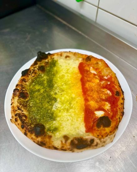Pizza rotonda su un piatto bianco con condimenti verdi, bianchi e rossi, appoggiata su un bancone della cucina.