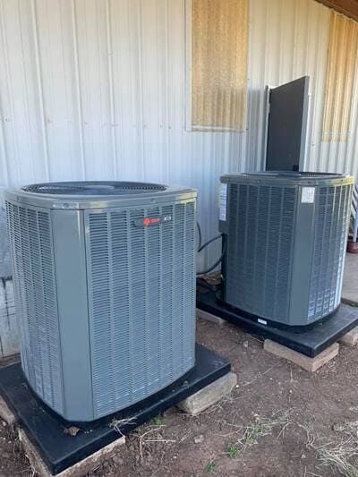 Two gray Trane HVAC outdoor units sitting on black plastic pads next to a white metal building.