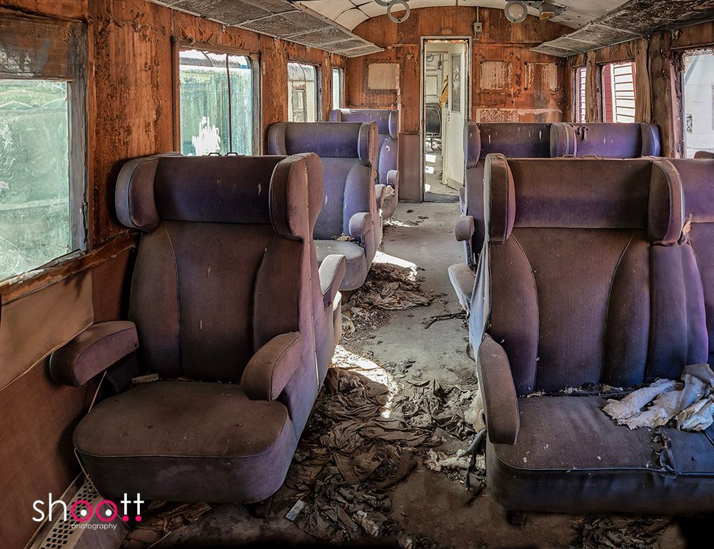 Een verlaten, vervallen treininterieur met versleten paarse stoelen en met puin bedekte vloeren onder een verweerd plafond.