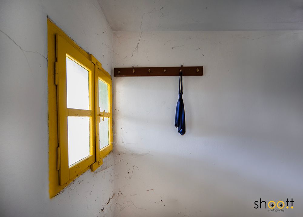 Een geel raam in een witte muur met een houten kapstok waaraan een blauwe stropdas hangt.