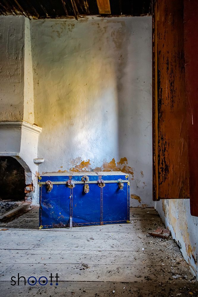 Een felblauwe opbergkist staat op een verweerde houten vloer in een rustieke verlaten kamer met afbladderende witte pleistermuren.