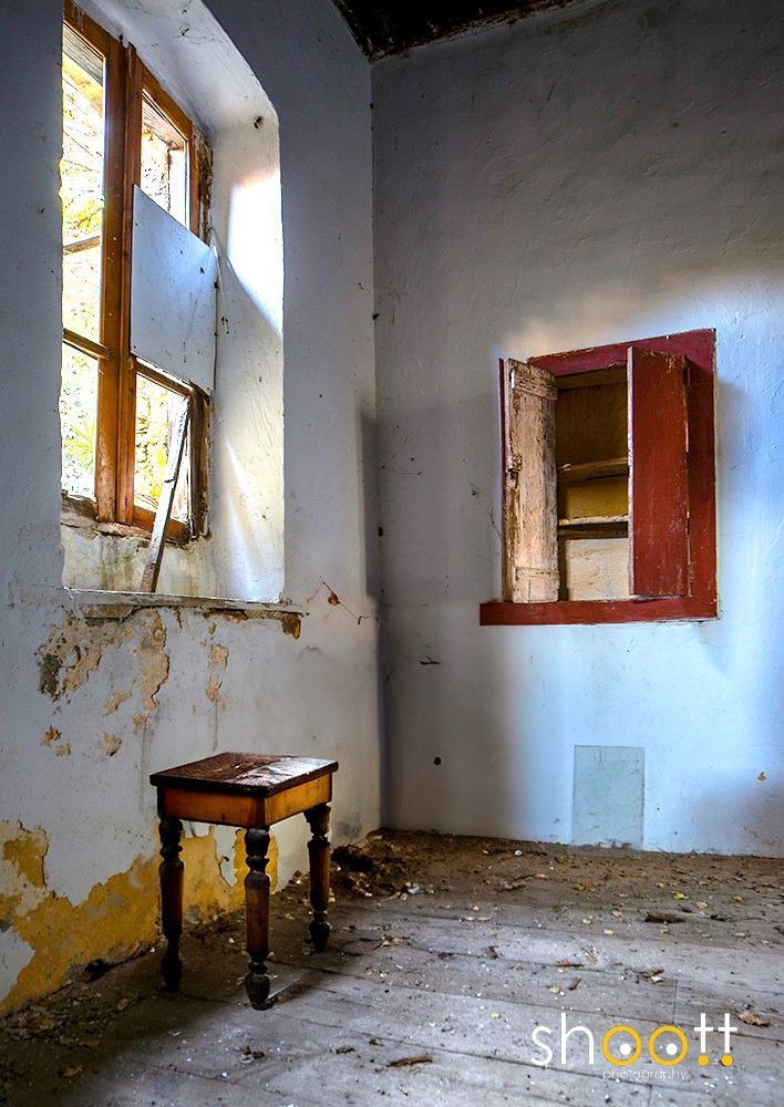 Een verlaten kamer met een klein houten krukje, een raam met een plank ervoor en een klein, rood omlijst wandkastje in een verlaten, vervallen huis.