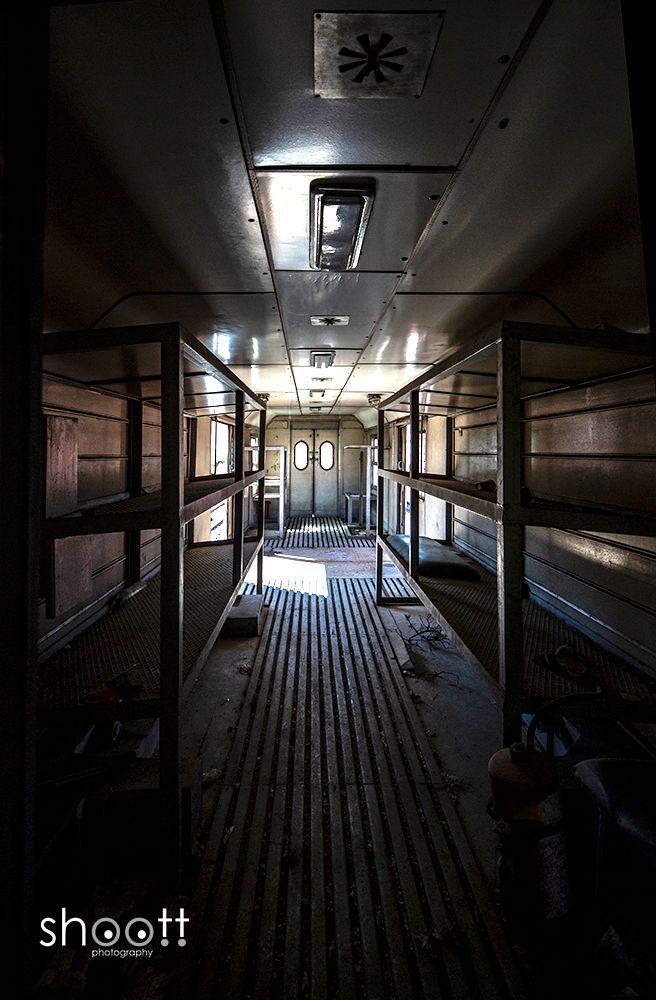 Een schemerig verlicht, smal treinstel met metalen schappen aan beide zijden en een lattenvloer die naar de deuren leidt.