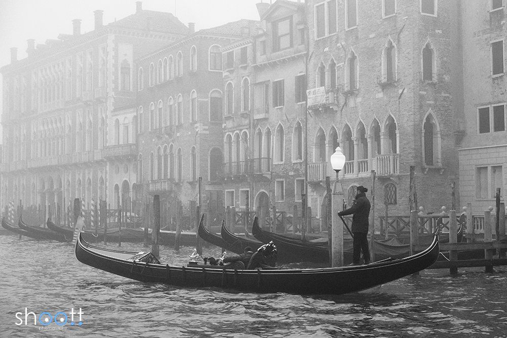 Een gondelier staat in een gondel in een mistig kanaal van Venetië, met historische gebouwen en aangemeerde boten op de achtergrond.