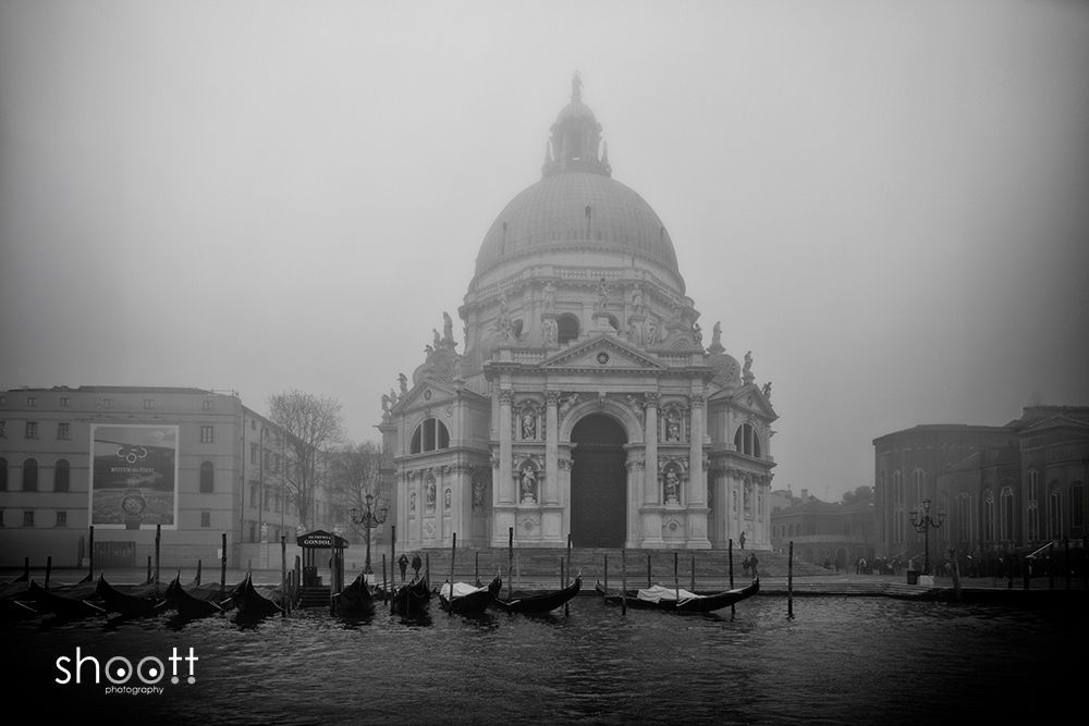 Een zwart-witfoto van de koepelvormige Santa Maria della Salute-kerk in Venetië, gehuld in dikke mist, met gondels op de voorgrond.