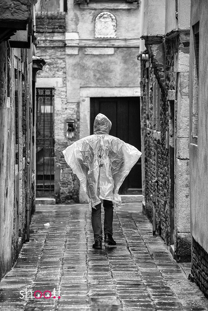 Een persoon in een doorschijnende regenponcho loopt weg door een smal, nat stenen steegje tussen verweerde bakstenen gebouwen.
