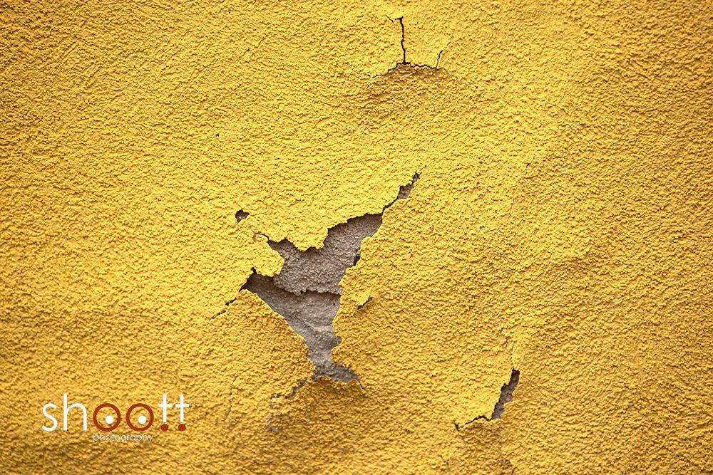 Een muur met een structuur, felgeel geverfd, waarvan een deel van de verf afbladdert en het ruwe grijze beton eronder zichtbaar maakt.