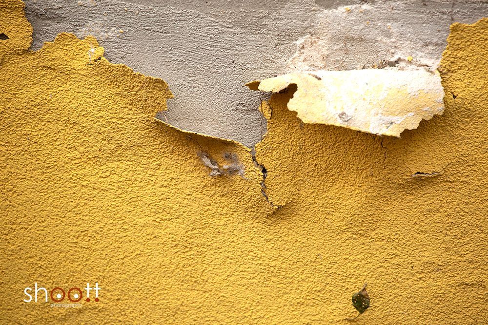 Een muur met een structuur in het geel, waar stukken pleisterwerk afbladderen en het grijze beton eronder zichtbaar wordt.