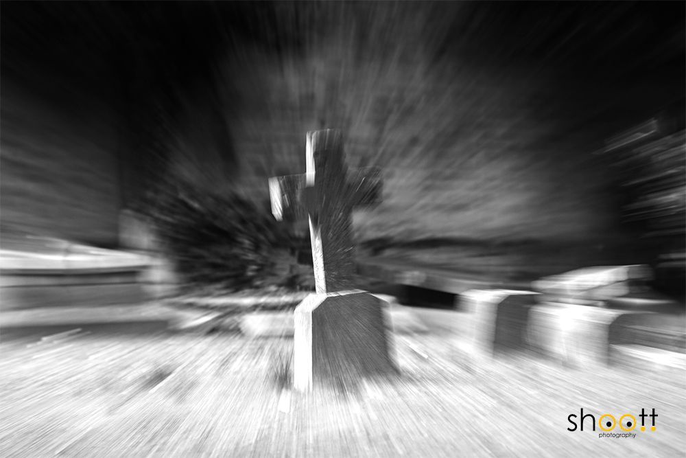 Een zwart-witfoto met radiale bewegingsonscherpte van een stenen kruis als grafmonument op een begraafplaats.