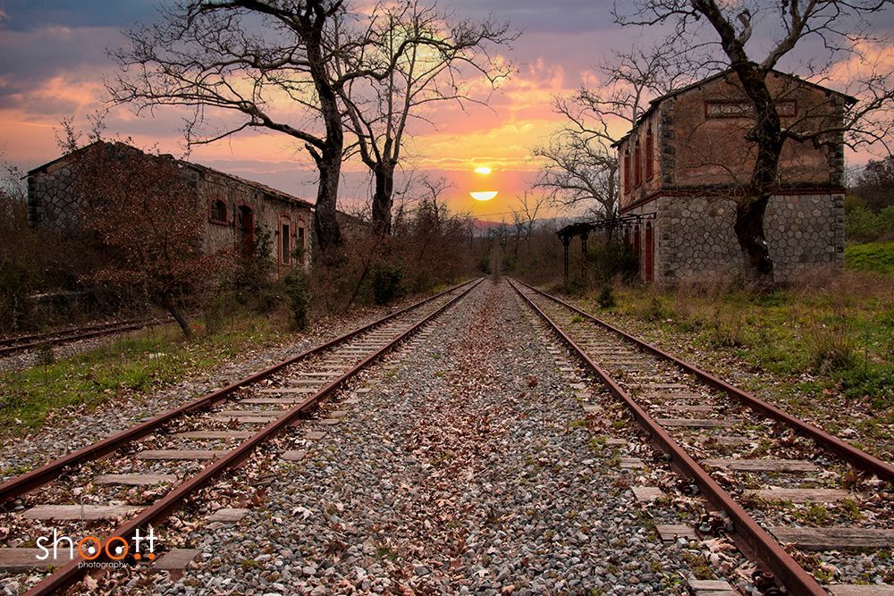 Verlaten stenen gebouwen flankeren overwoekerde spoorlijnen die leiden naar een zonsondergang met een hemel vol warme, gouden tinten.