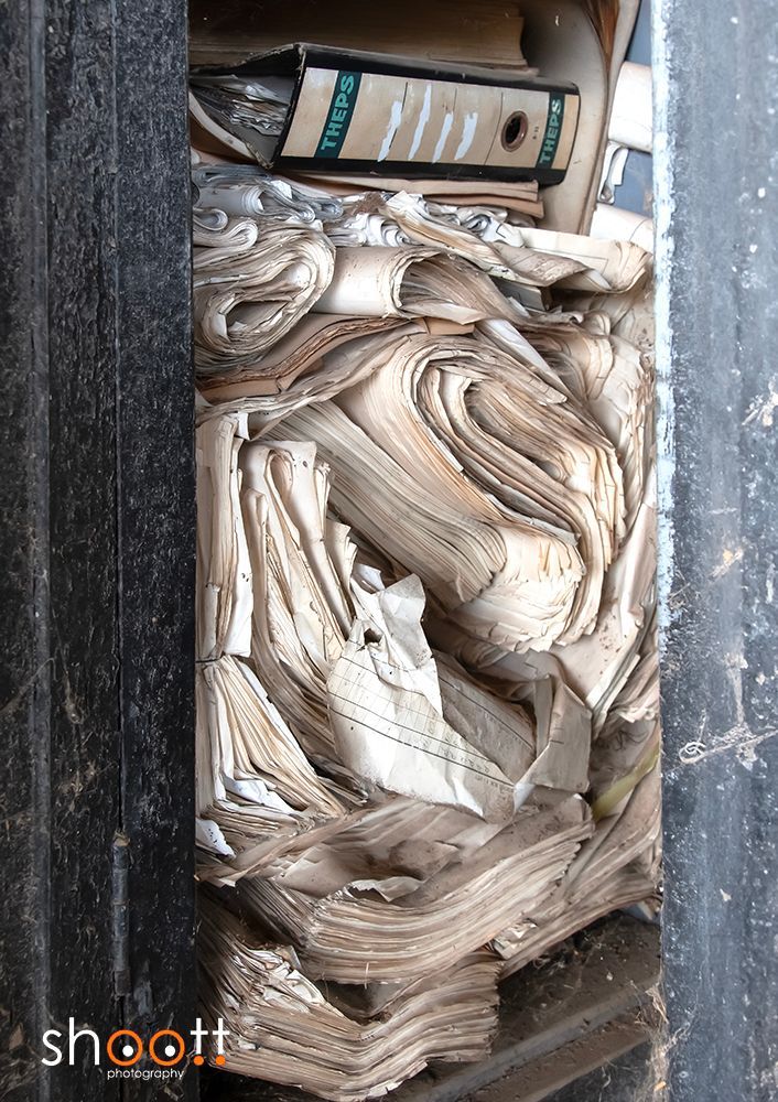 Een close-up van een rommelige plank met een donkere map bovenaan en stapels versleten, opgevouwen papieren dossiers die de ruimte vullen.