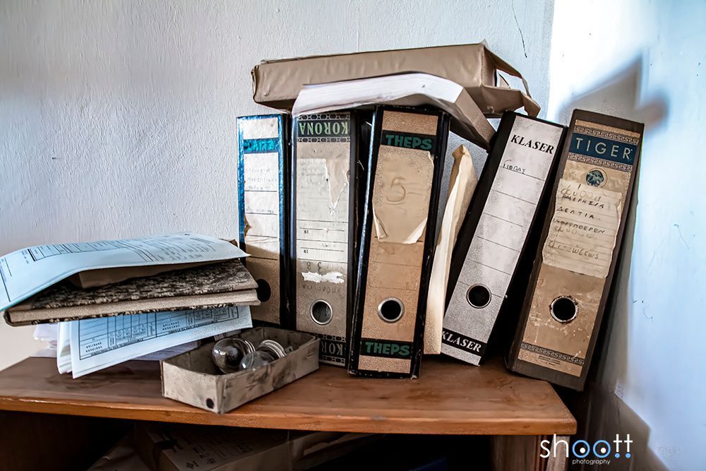 Op een houten plank tegen een hoekmuur in een schemerig verlichte kamer staan ​​diverse versleten, van etiketten voorziene ordners en een stapel papieren.