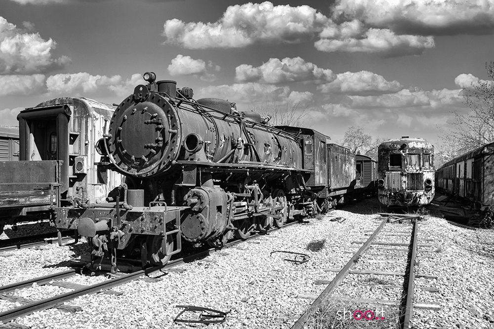 Een zwart-witfoto van een oude, verweerde stoomlocomotief die op roestige spoorrails in een rangeerterrein staat.