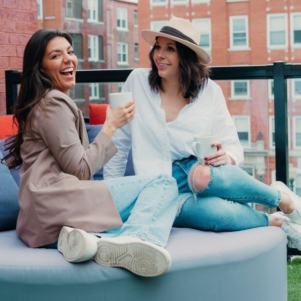 Two women are sitting on a couch drinking coffee and laughing.