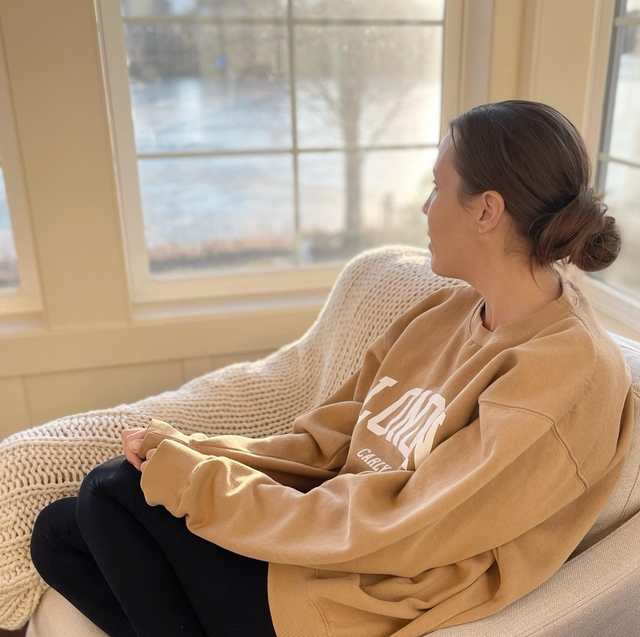 Amy sitting with her legs tucked up in a cream chair looking out a window on a lake and hands folded in her lap. Wearing a brown London sweatshirt and black leggings with a messy low bun. 