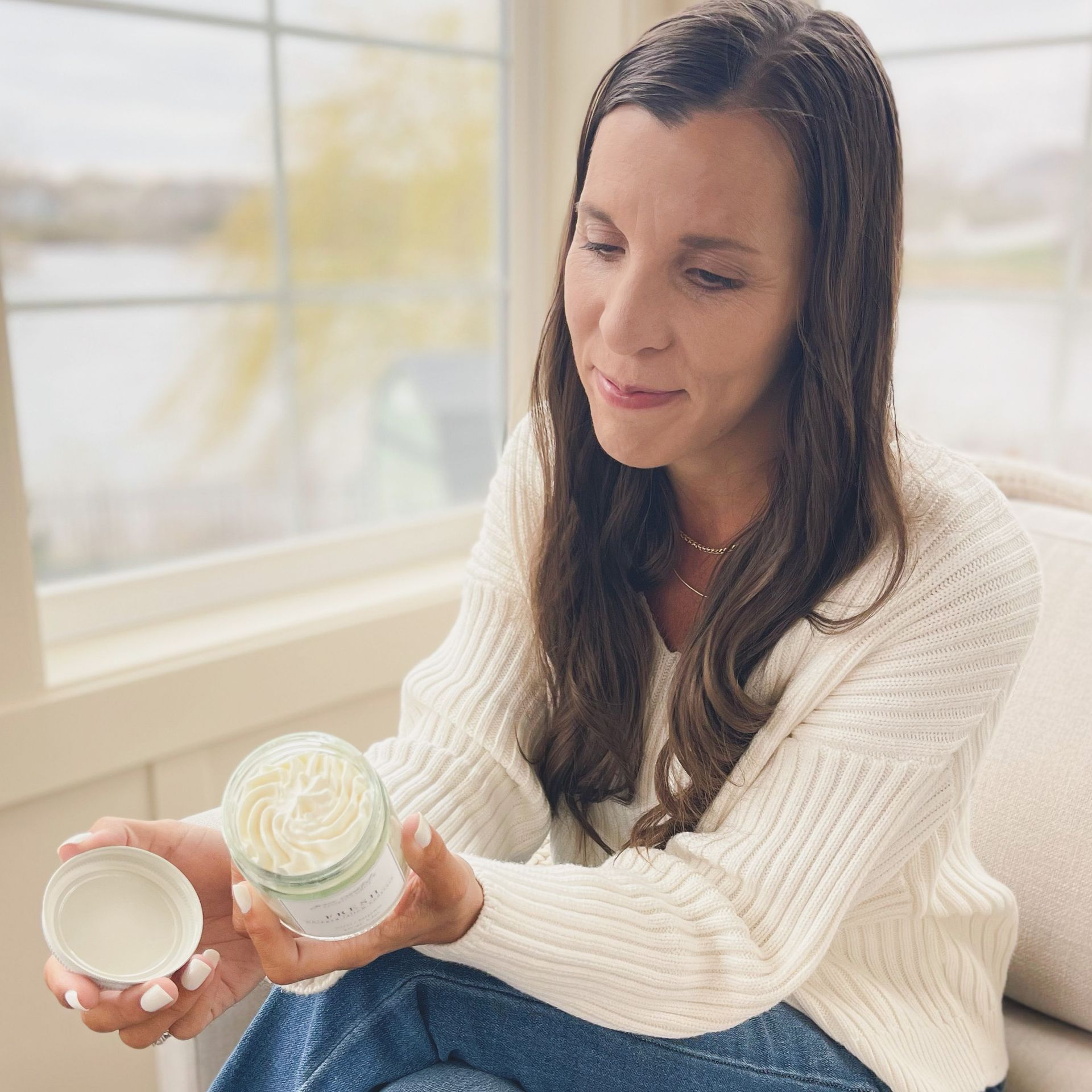 Amy sitting in a cozy white chair with windows behind her looking out to a lake. She is smiling off to the side with her hands in her lap and her legs tucked up on the chair.