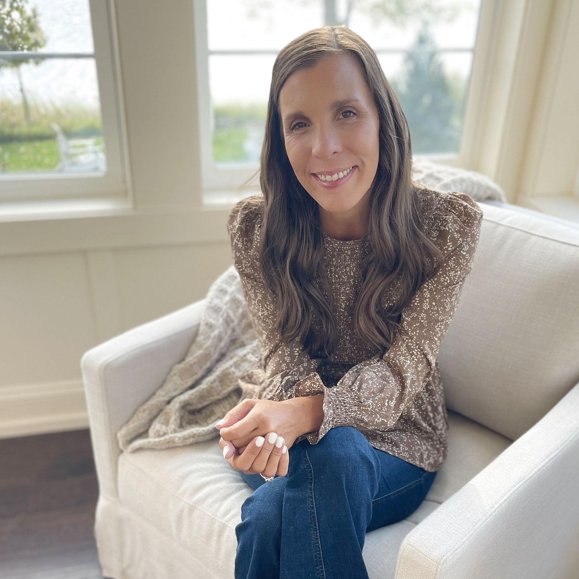 Amy sitting on chair - legs crossed and hands folded on her lap leaning forward with a smile. Wearing a brown dress shirt with dark blue jeans. Windows behind her overlooks a lake with green grass.