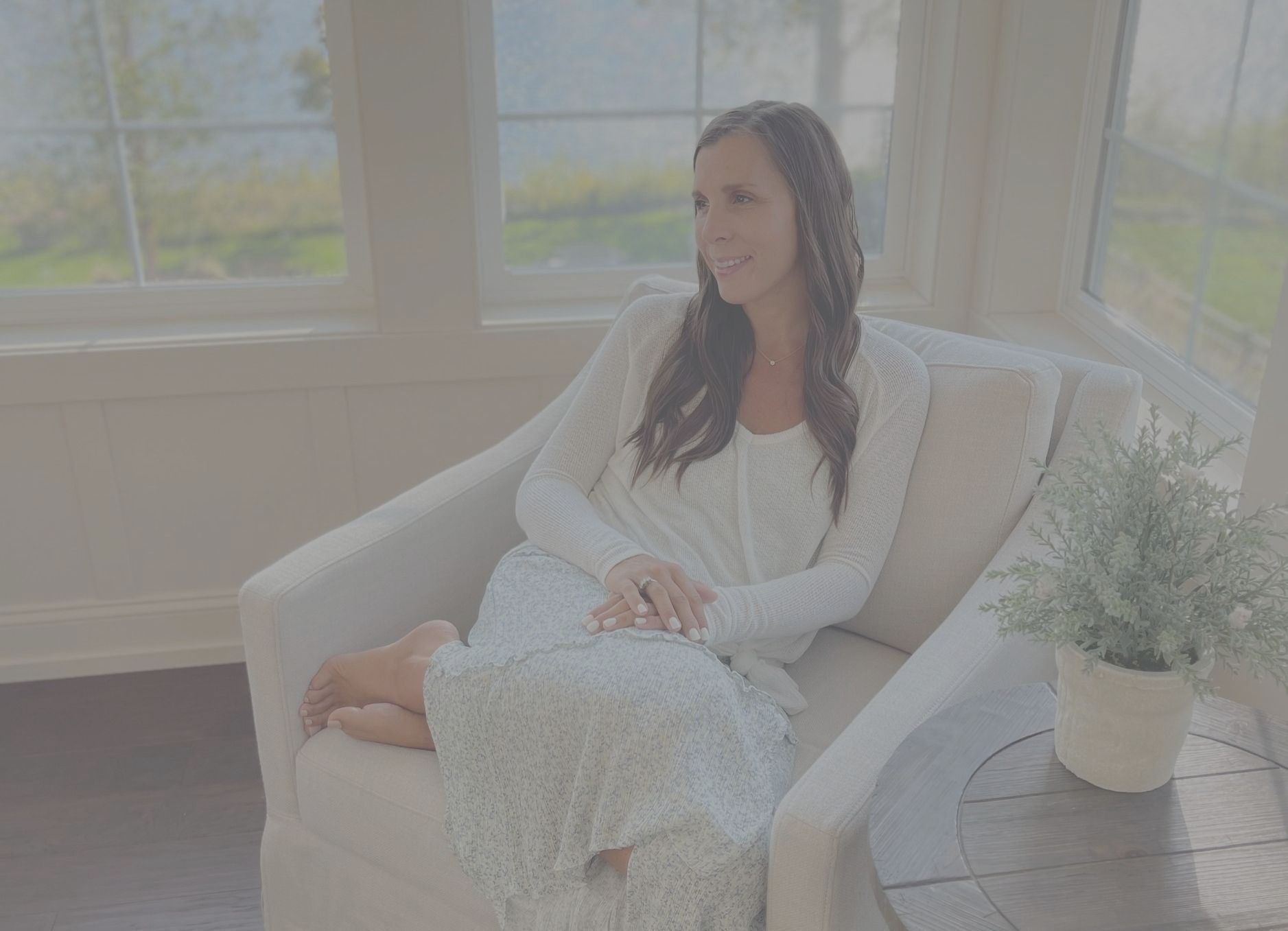 Contrasted photo of Amy looking off in the distance smiling. White top  and blue flower skirt on bottom sitting in cozy white chair with windows and a lake in the background. White nail polish. Happy photo.