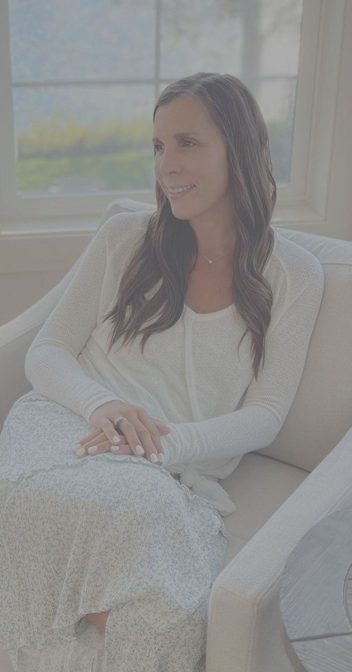 Contrasted photo of Amy looking off in the distance smiling with bible and journal in lap. White top  and blue flower skirt on bottom sitting in chair next to fireplace. Hair down and flowy beach curls. Single diamond necklace on. White nail polish. Happy photo.