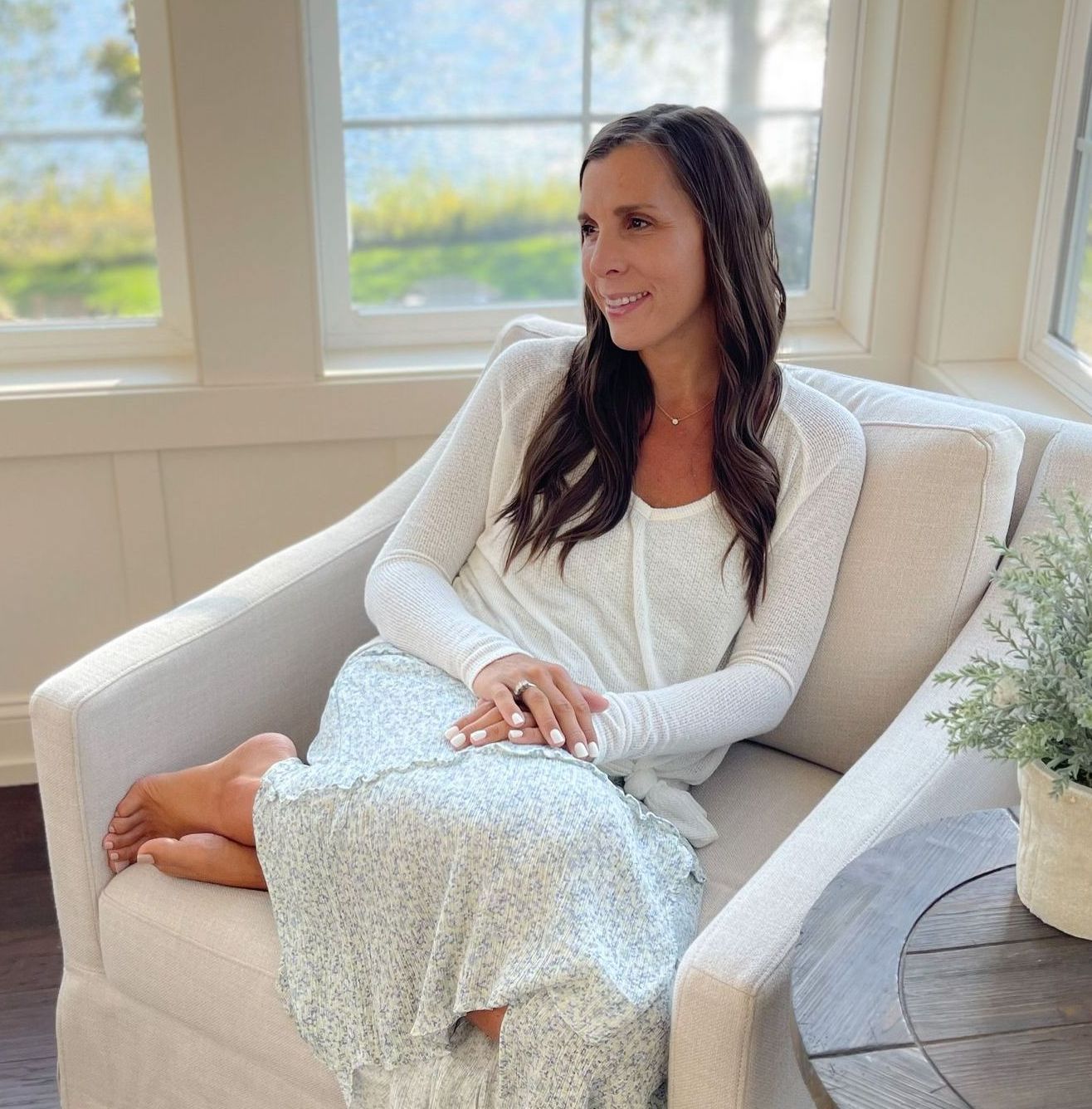 Side image of Amy sitting in chair with hair down and wavy. Bible and journal on lap with legs crossed. Smiling in blue flower skirt and white long sleep top