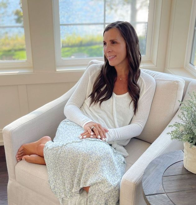 Side image of Amy sitting in chair with hair down and wavy. Bible and journal on lap with legs crossed. Smiling in blue flower skirt and white long sleep top