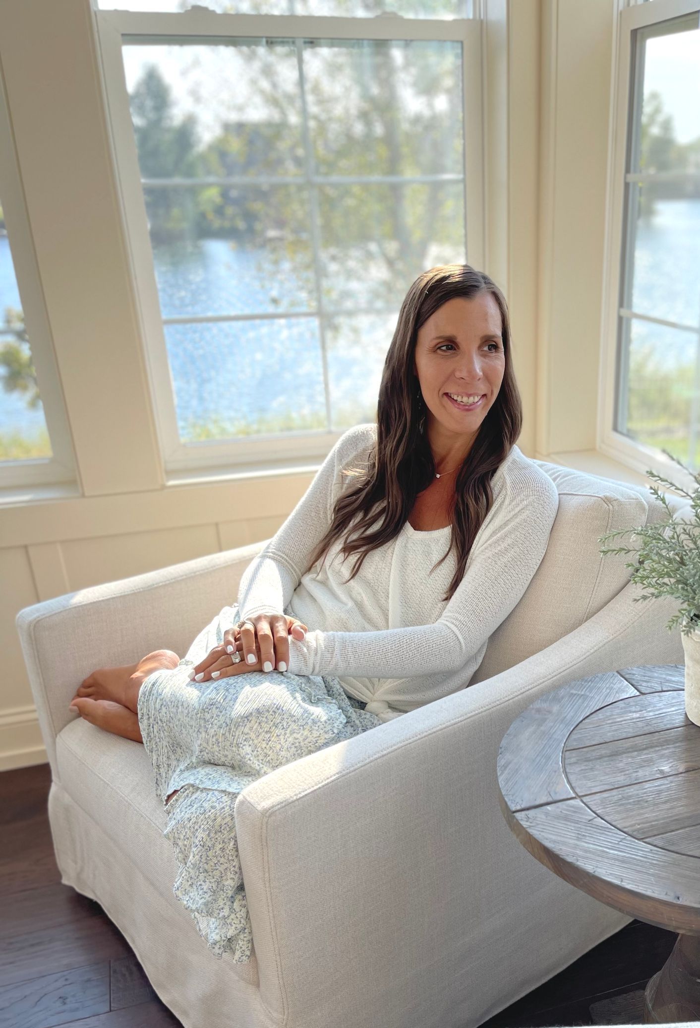 Bright photo of Amy sitting in creamy pad chair with wooden back and arms. Legs pulled up on chair. Hair down and wavy curls. Smiling big with bible and journal in lap. Looking  directly at camera.  Same outfit as above with white long sleeve top and blue and white flowy skirt. White sandals on. White nail polish on hands and feet.