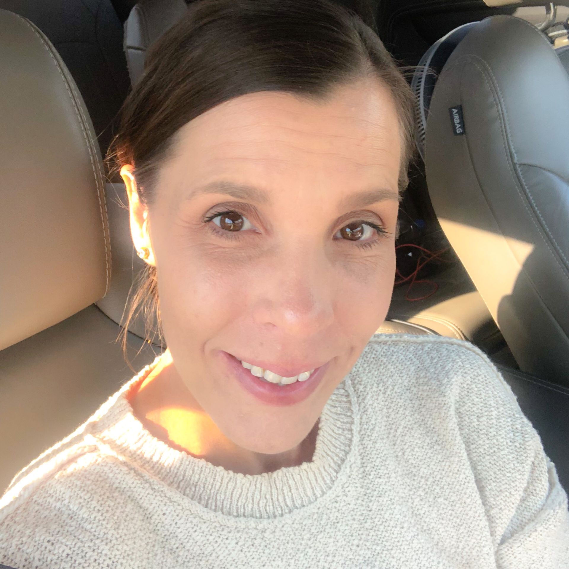 Amy taking  a selfie in the car - full smile with the sun hitting her.  Wearing a beige sweater.