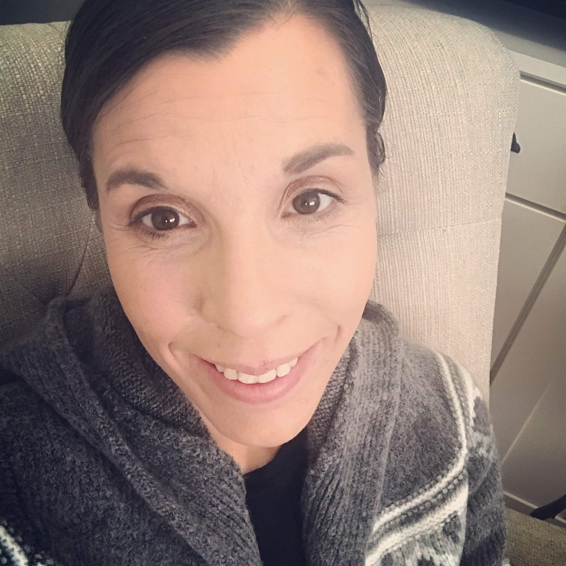 Amy sitting in cream chair with big smile. Hair pulled back. Open gray sweater on with black shirt