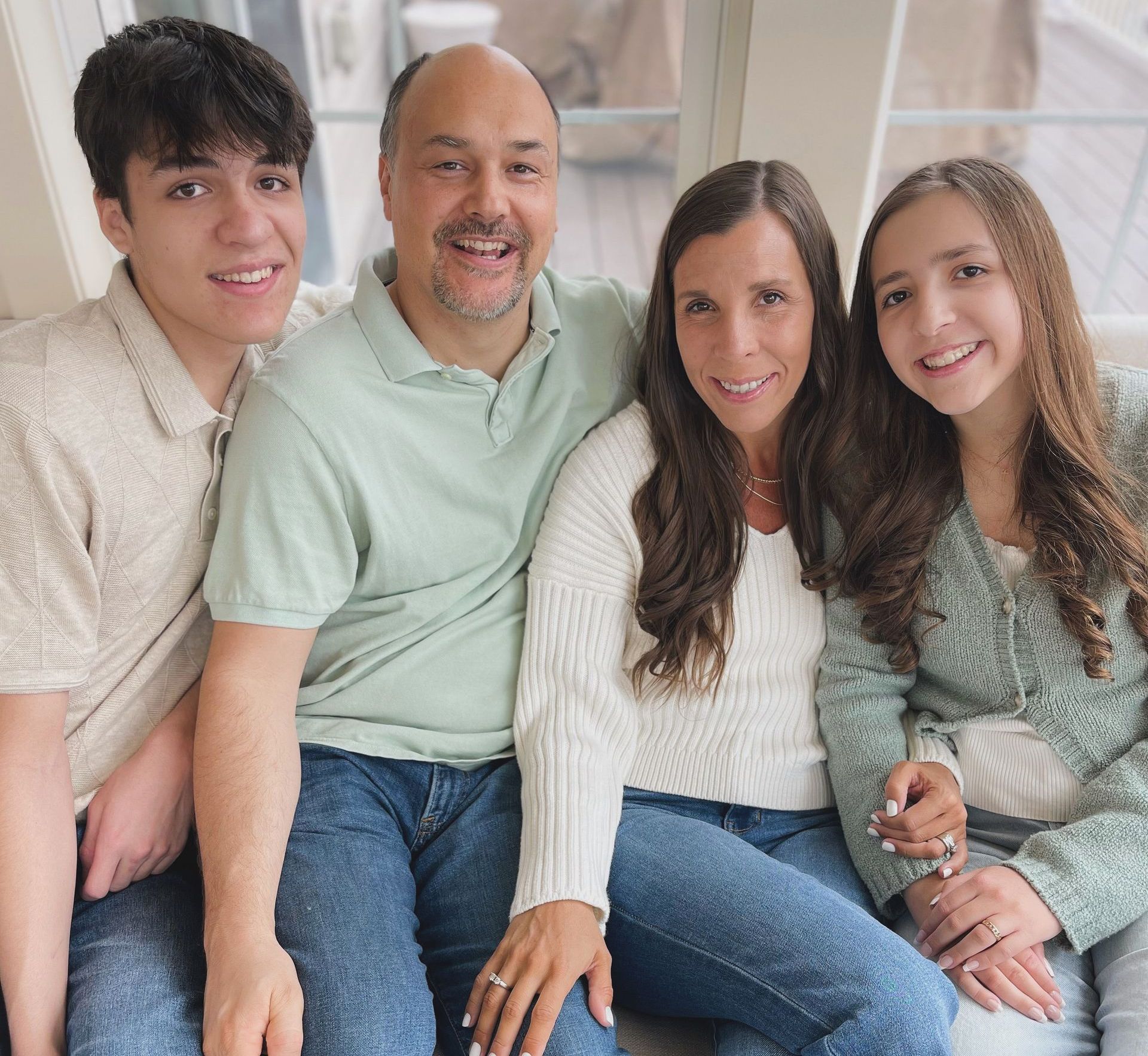 Photo of Randy, Mason, Ella and Randy next to fireplace.  Amy sitting in chair while the rest kneel around her. All smiling big wearing blue,white, and creamy tan colors. Looking directly at camera. Grateful sign behind them.