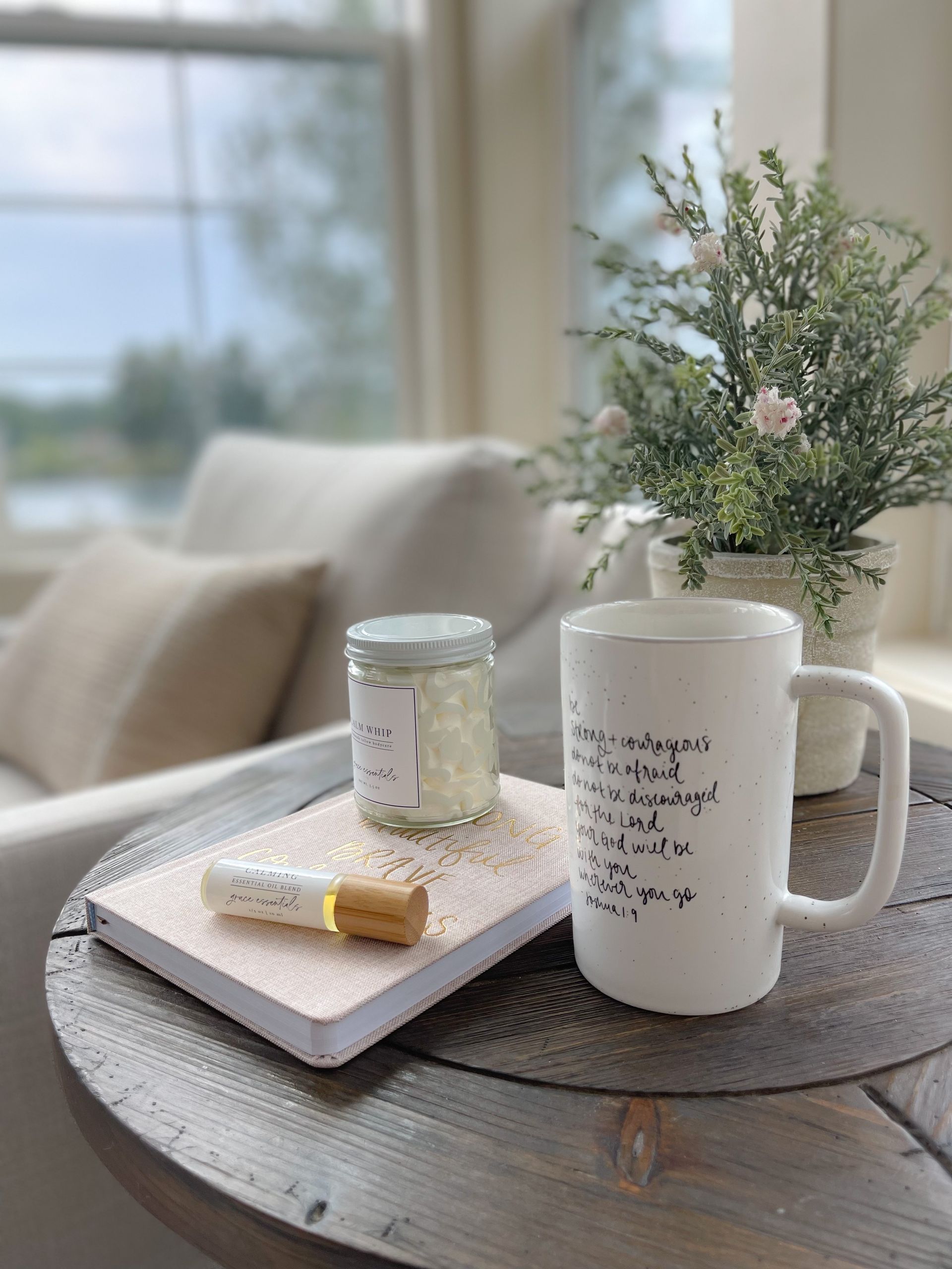 Wooden end table with blurry chair and window in the background. On the table are Grace Essentials products - bible verse mug, essential oil roller blend, whipped tallow and journal.