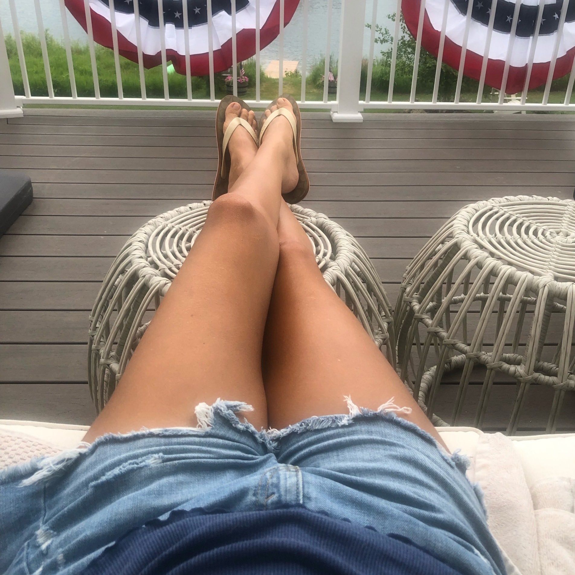 Amy outside on deck legs up on stool cut off denim shorts.  Lake in background with american  flags