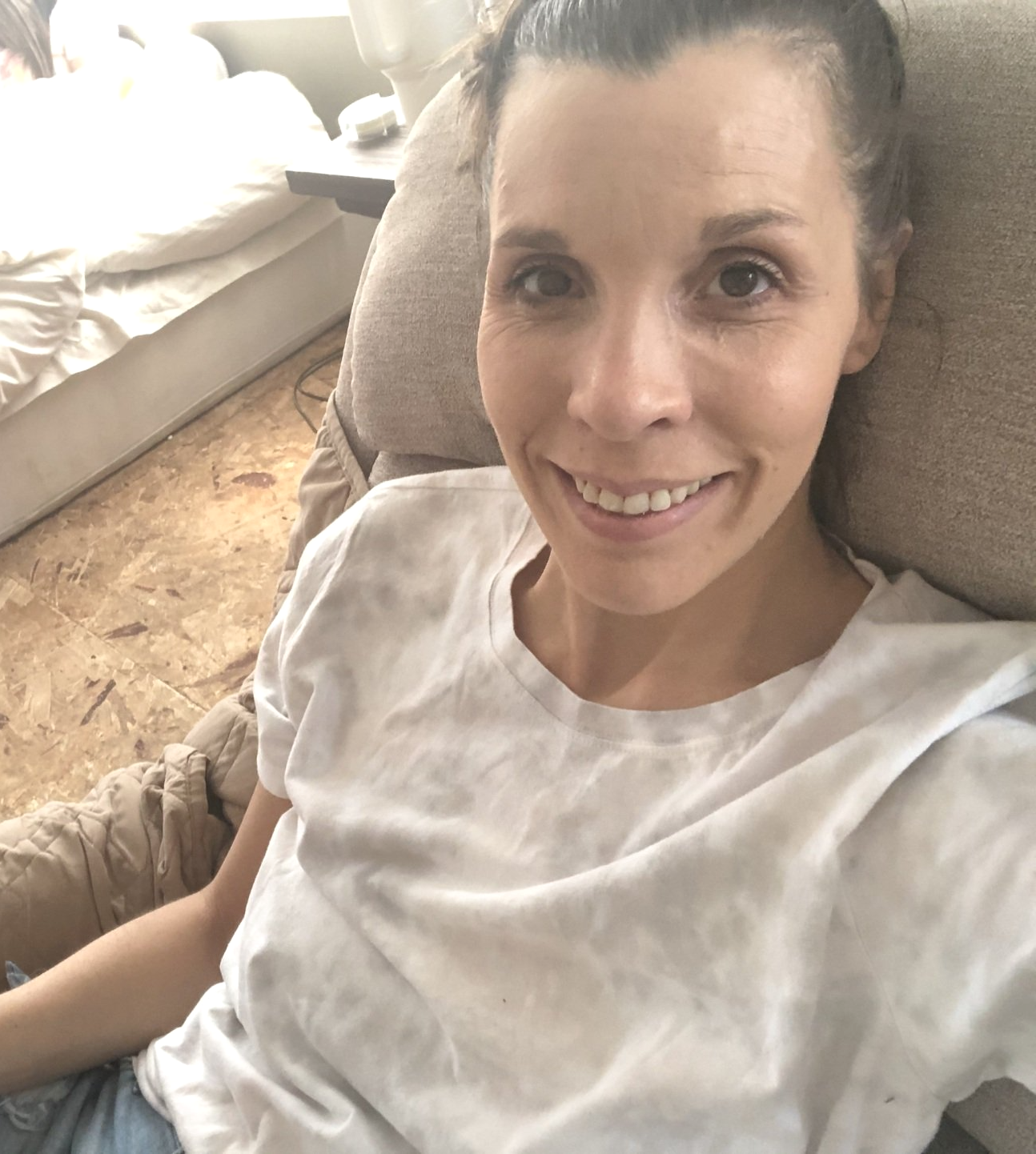 Amy smiling in recliner. Hair pulled back.  Looking very thin and sick colored skin. No carpet flooring and bed on floor behind her