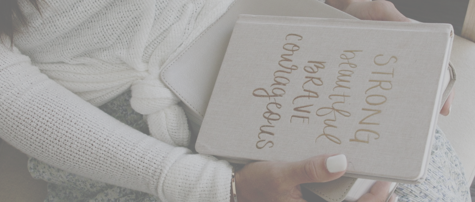 Close up of tan Journal on top of bible sitting on Amy's lap that says  strong beautiful brave courageous.  Contrasted and creamy photo. Amy wearing white long sleeve shirt and blue and white flowy skirt. White painted nails.